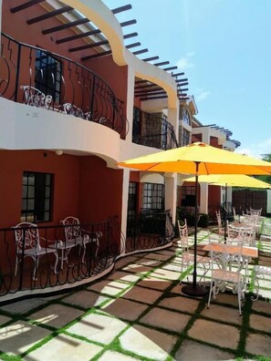 Un patio con mesas al aire libre, un paraguas amarillo y un edificio con balcones.