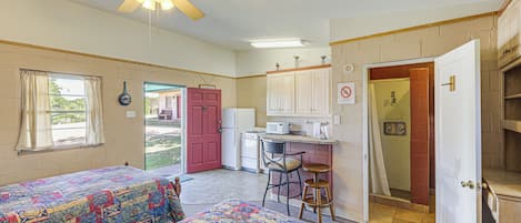 Una habitación con dos camas, kitchenette, ventilador de techo y puerta al baño.