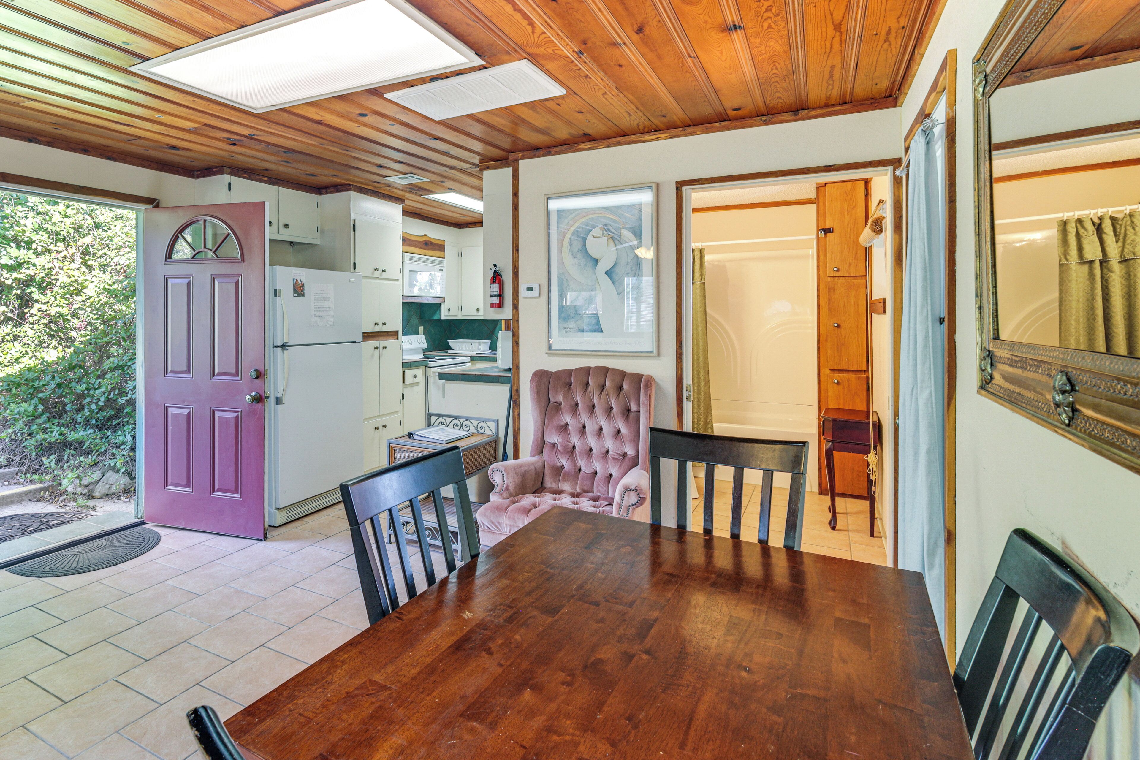 A wooden ceiling, a dining table with chairs, a pink tufted chair, a kitchen with white cabinets, and a door with a purple frame.