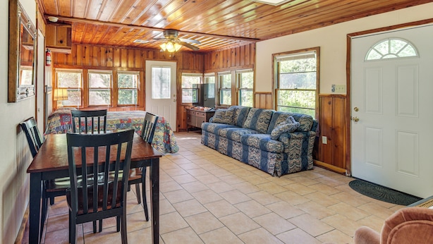 A spacious living area with a wooden ceiling, a sofa, a dining table with chairs, and large windows.