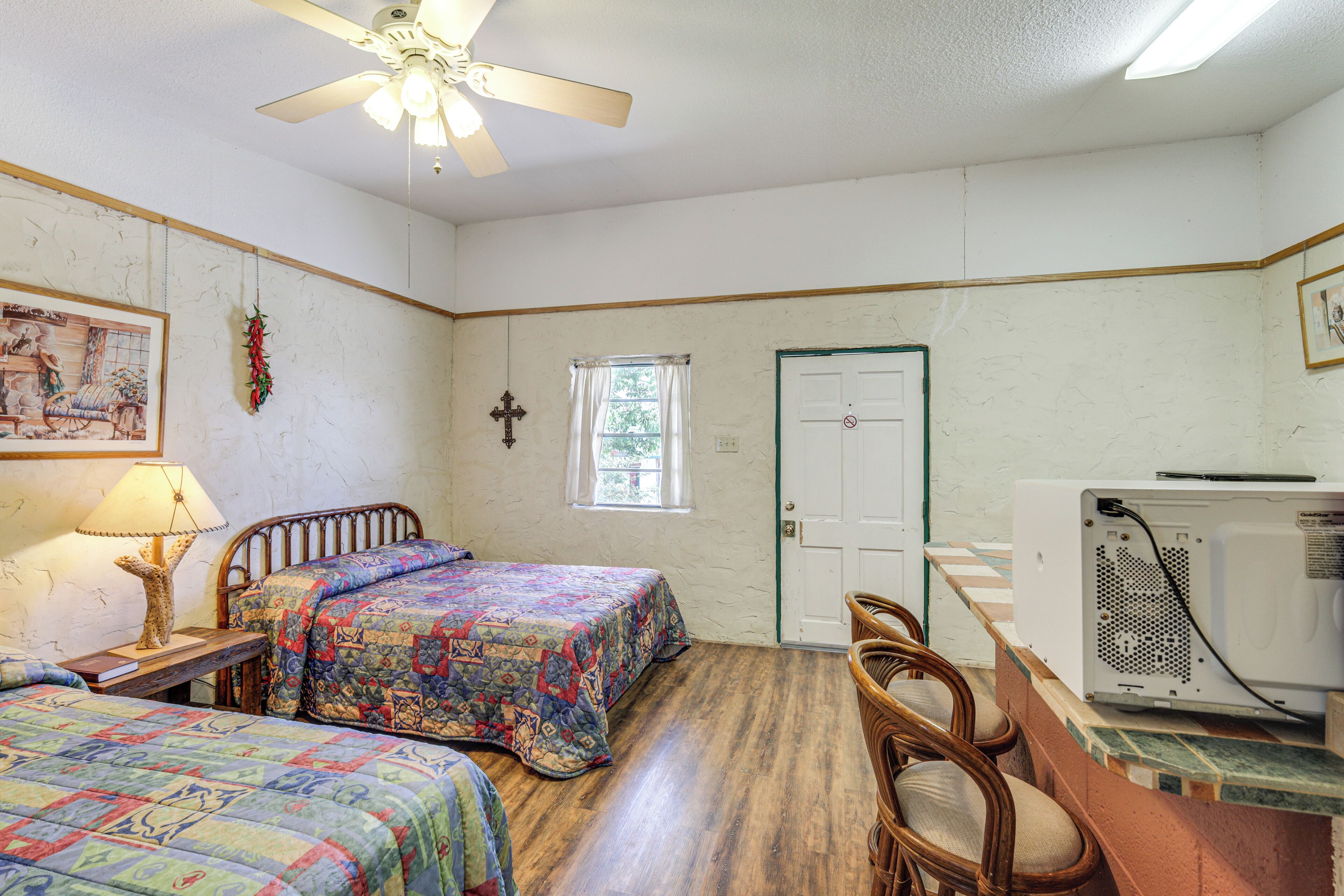 A room with two beds, a ceiling fan, a window with curtains, a door, and a wall-mounted heater.