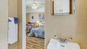 Habitación de hotel con piso de madera, dos camas con ropa de cama colorida, un ventilador de techo, un lavamanos blanco y un espejo sobre el lavamanos.