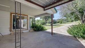 Une véranda couverte avec un plancher en béton, une échelle en métal et une vue sur un espace verdoyant avec des arbres et un sentier.