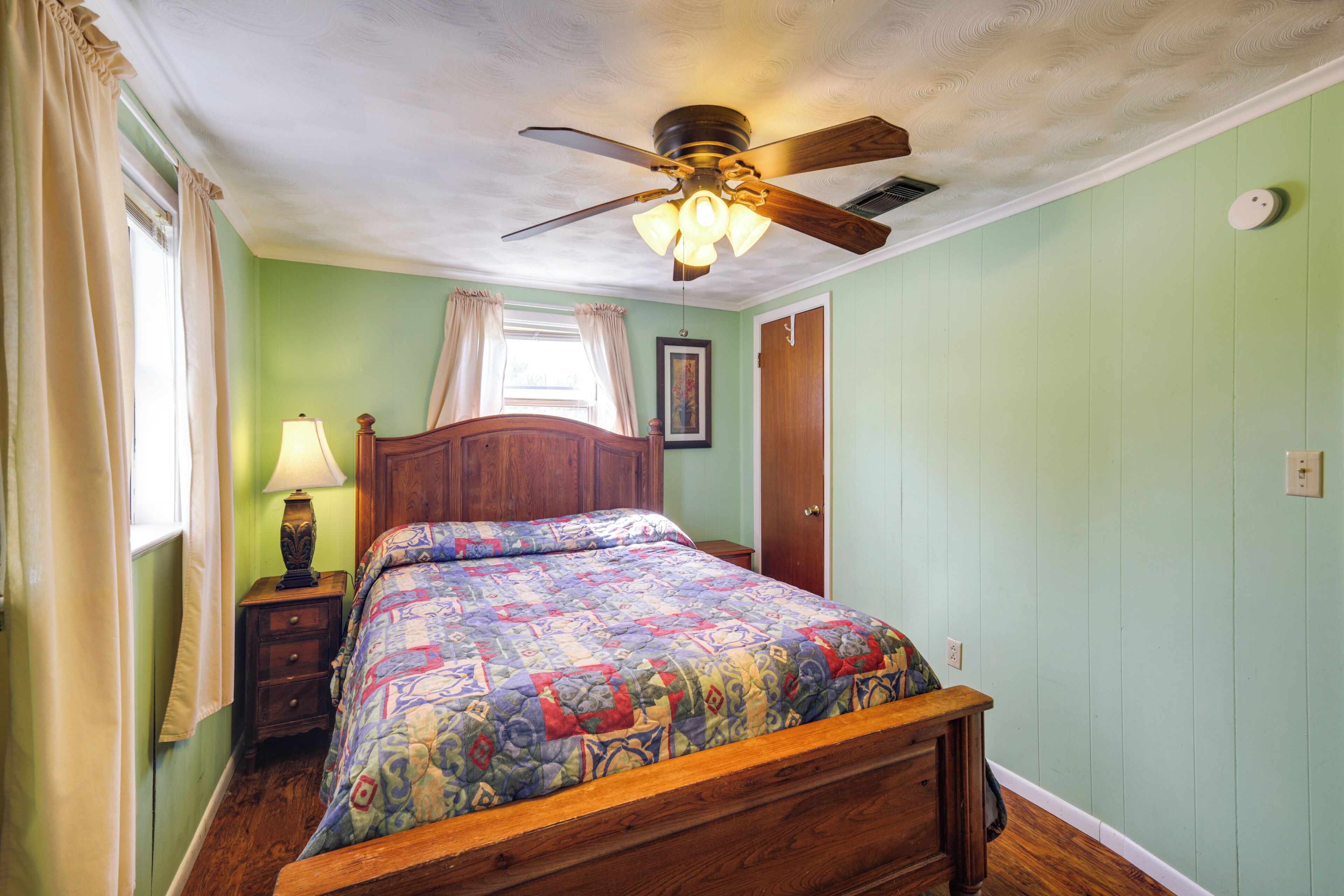 A bedroom with a wooden bed, a nightstand with a lamp, a ceiling fan, a window with curtains, and a door.