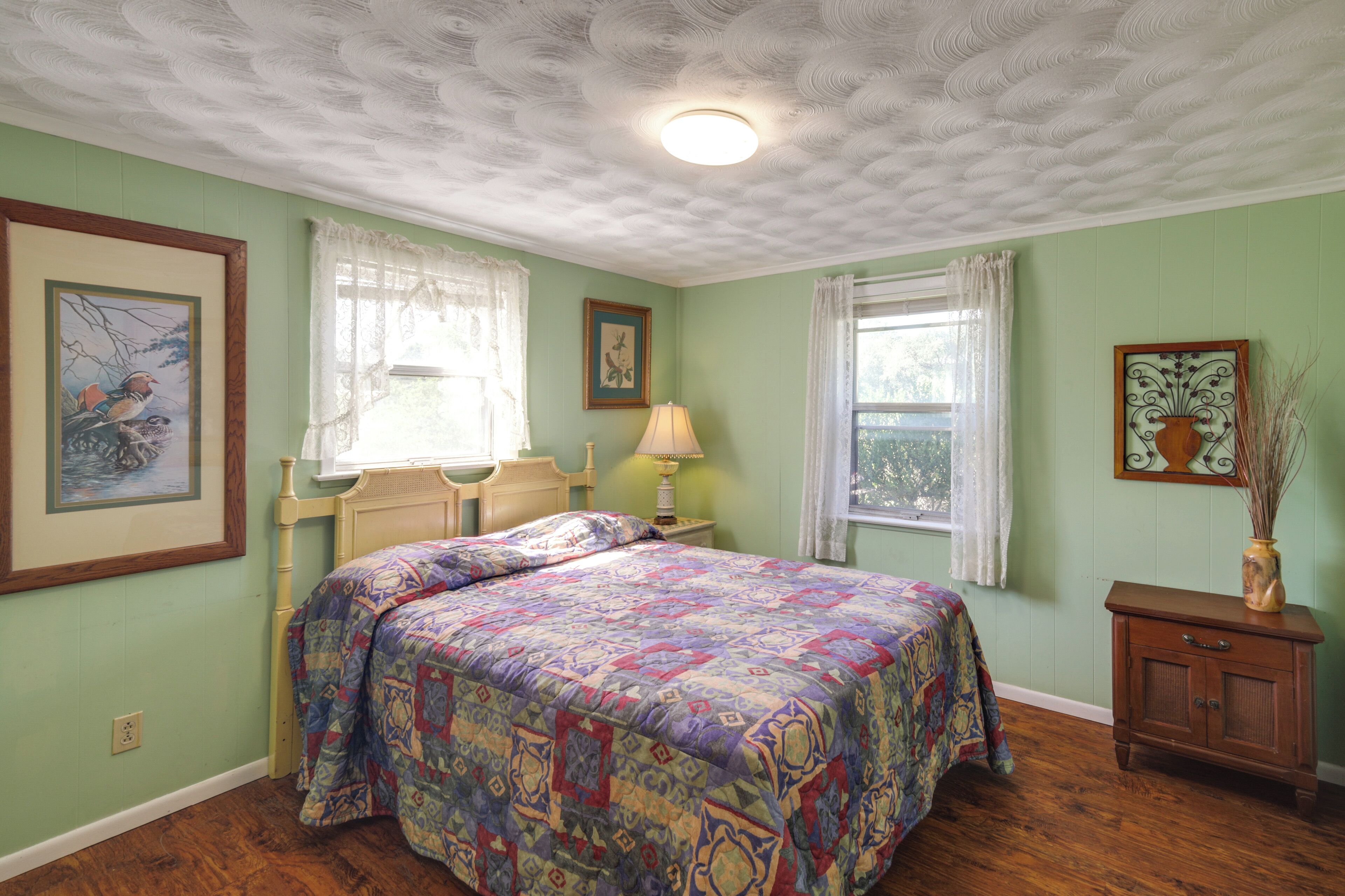 A bedroom with a bed, a nightstand, a dresser, and two windows with curtains.