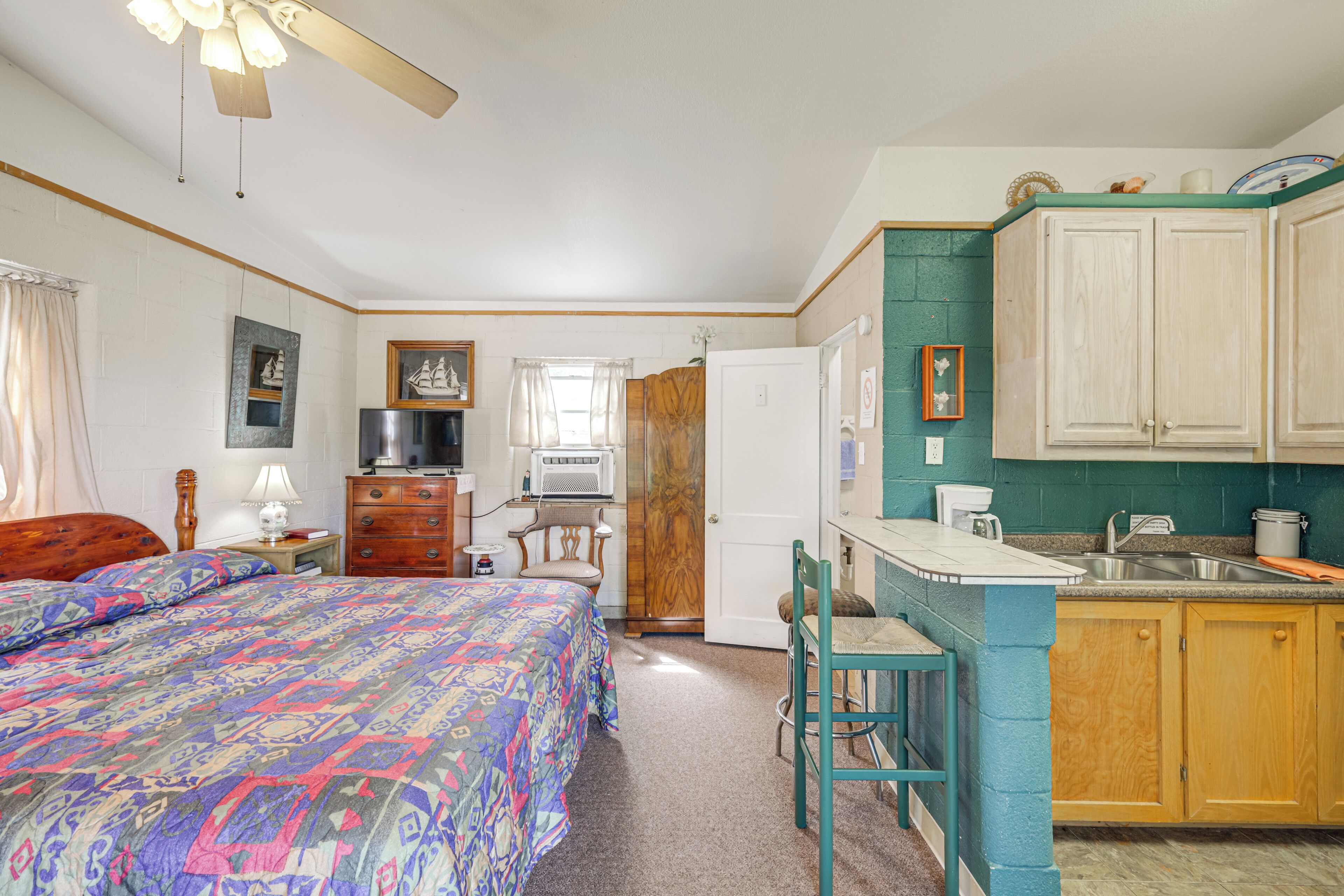 A room with a bed, a desk, a chair, a kitchenette, and a door leading to another room.