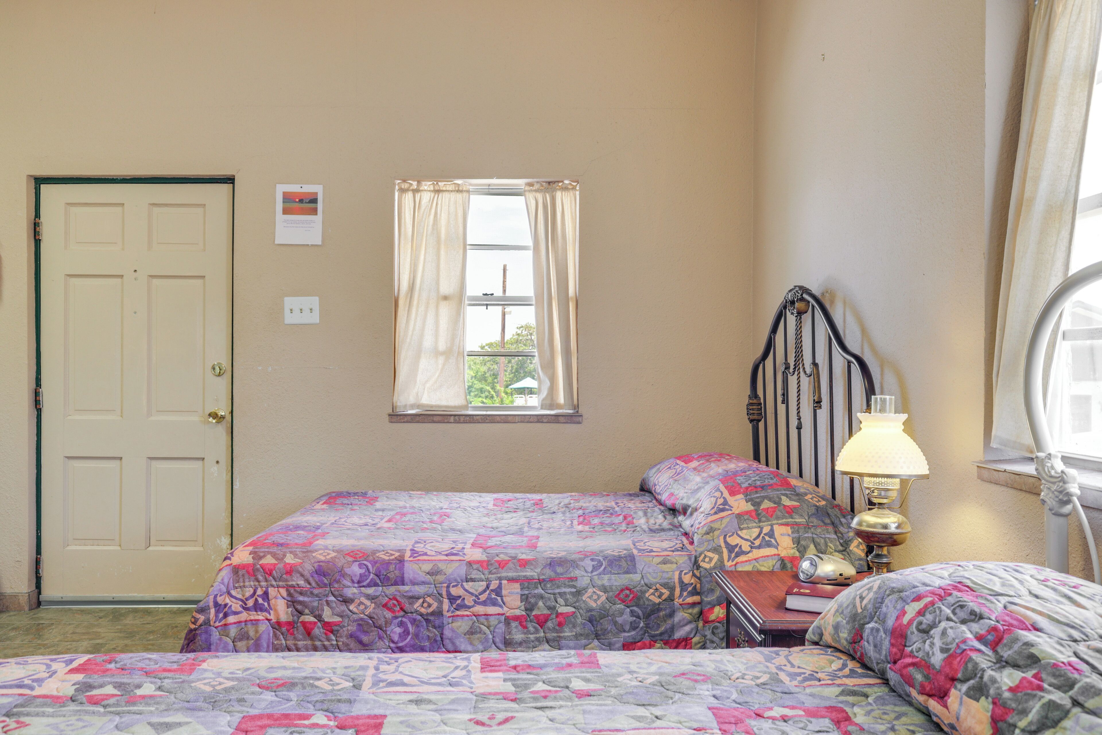 Un dormitorio con dos camas, una ventana con cortinas, una lámpara de noche y una puerta.