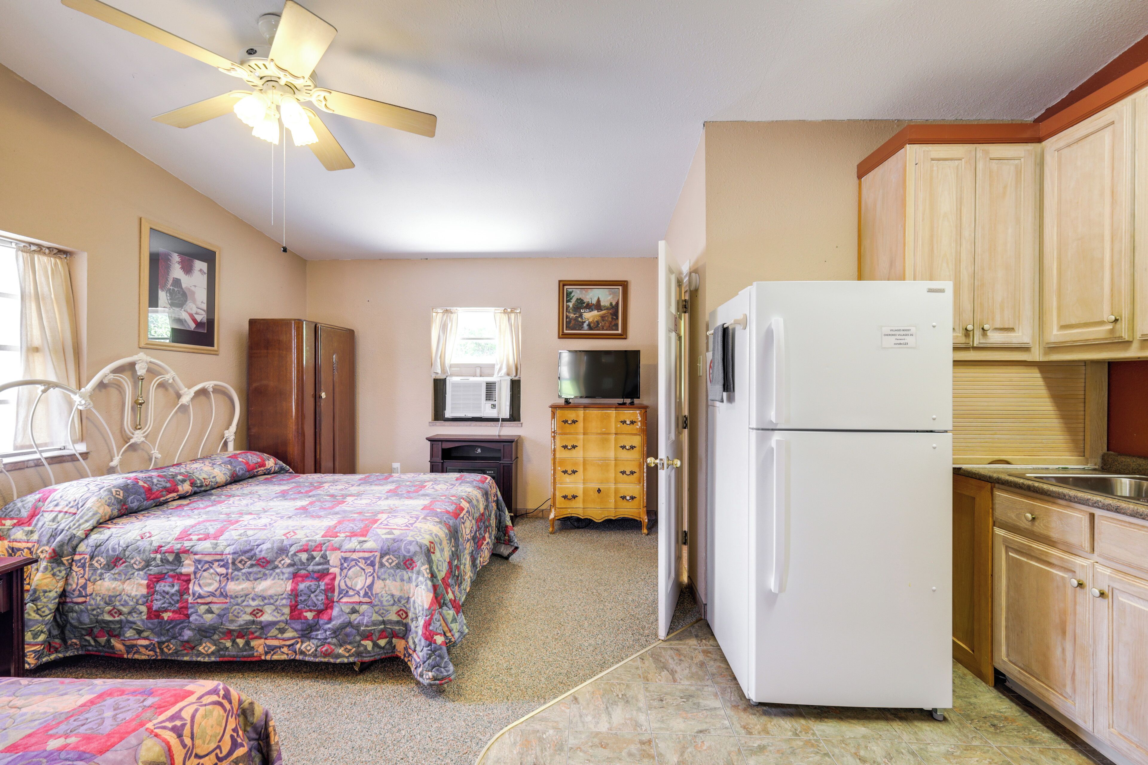 A compact room with a bed, a refrigerator, a television, and a ceiling fan.