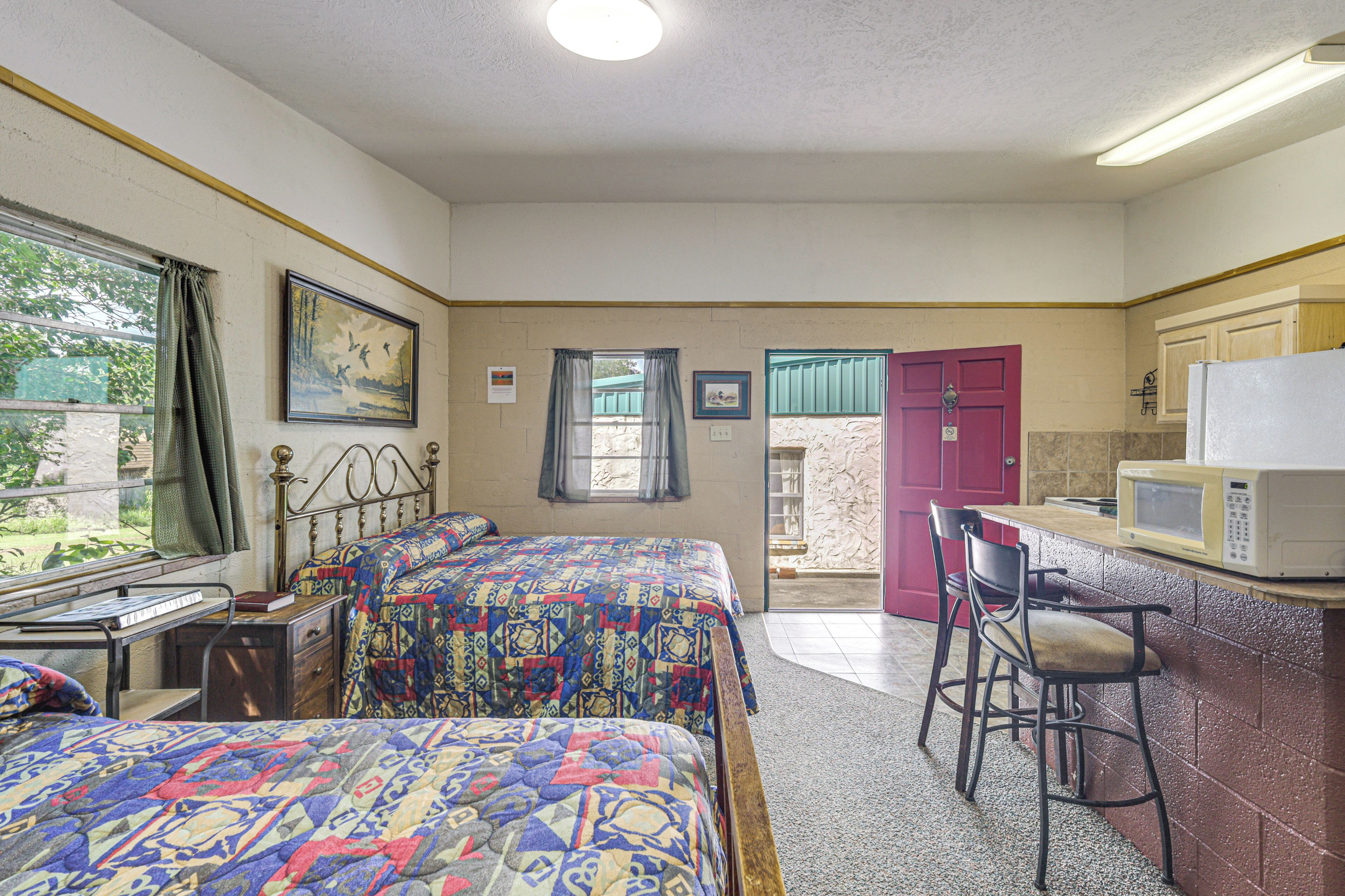 A bedroom with two beds, a desk, a chair, a microwave, and a red door leading to a bathroom.