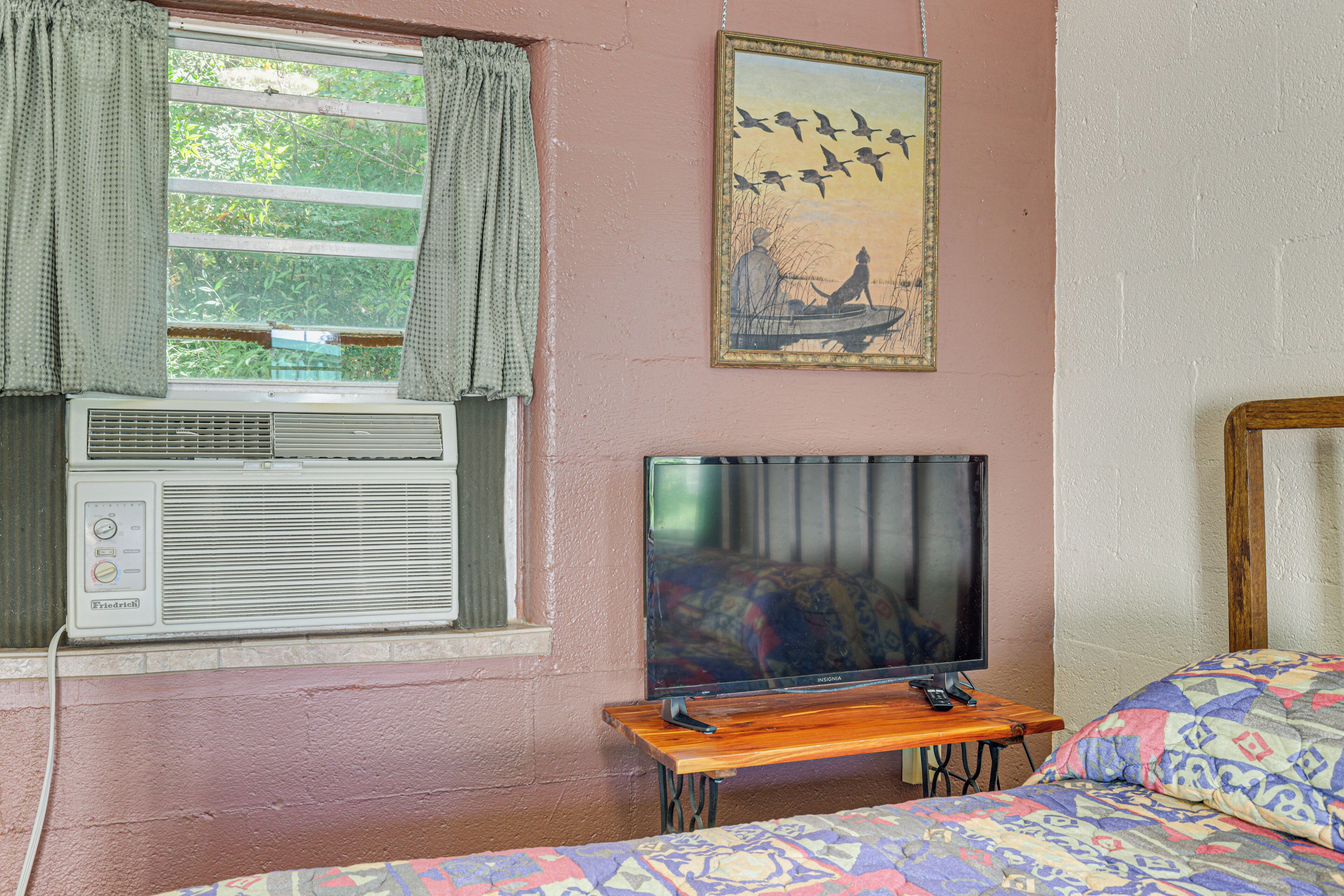 A room with a bed, a television on a wooden stand, an air conditioning unit, and a painting on the wall.