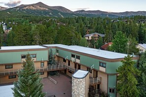 A multi-story building with a flat roof, surrounded by dense forest and mountains in the background.
