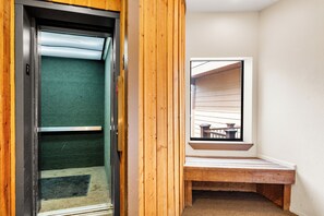 Un pasillo estrecho con un banco de madera, una ventana que da a muebles de exterior y una puerta con un felpudo verde.
