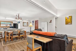 Ein modernes Wohnzimmer mit einer Couch, einem Esstisch und Stühlen. Es gibt eine Küchenzeile mit einem Kühlschrank und einem Fenster.