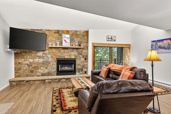 A living room with a stone fireplace, a leather sofa, a flat-screen TV, and a large window.