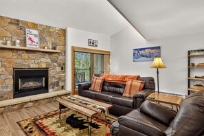 A cozy living room with a stone fireplace, leather sofa, and a view of the outdoors. - Top-Floor Ski-In Condo | Walk to Lifts & Main St (Breckenridge)