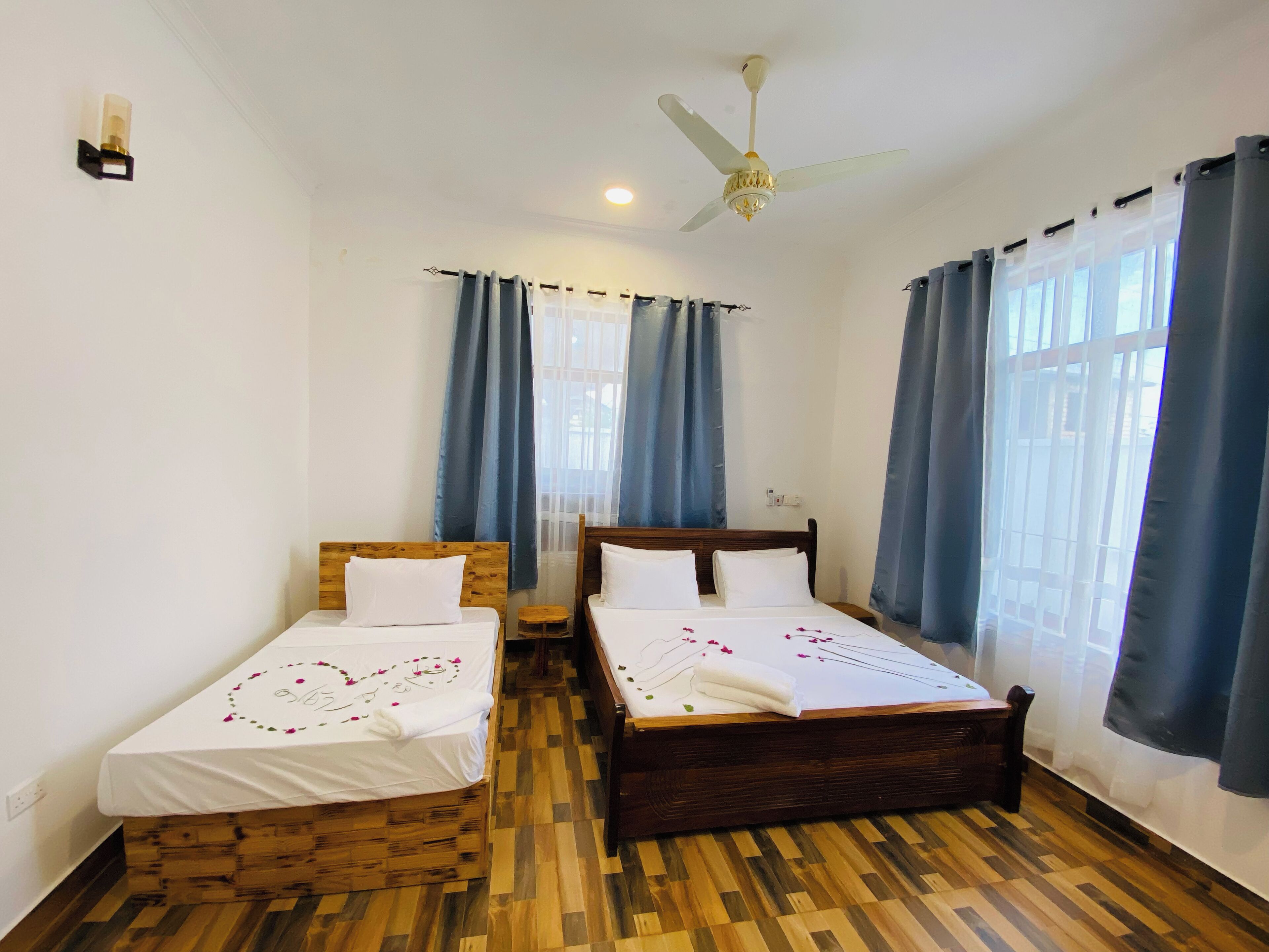 Una habitación con dos camas de madera, un ventilador de techo y ventanas grandes con cortinas azules.