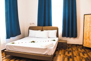 Una cama bien tendida con sábanas blancas y un cabecero marrón, una mesita de noche pequeña con un cajón, y suelo de madera.