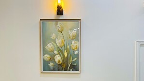 A wooden table with a bench in a room with a plain white wall and a framed painting of flowers.