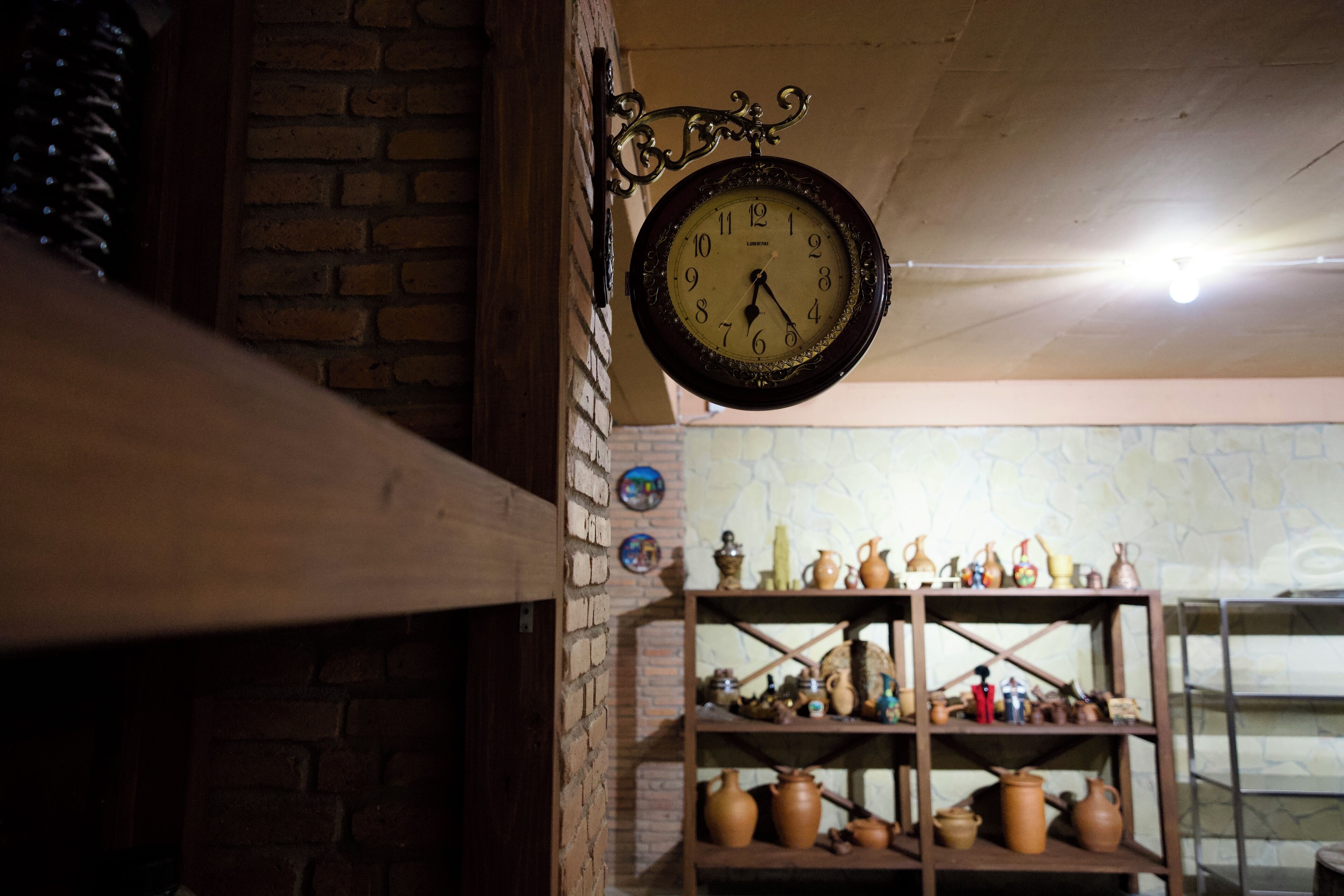 A rustic brick wall with a hanging wall-mounted clock and shelves displaying pottery.
