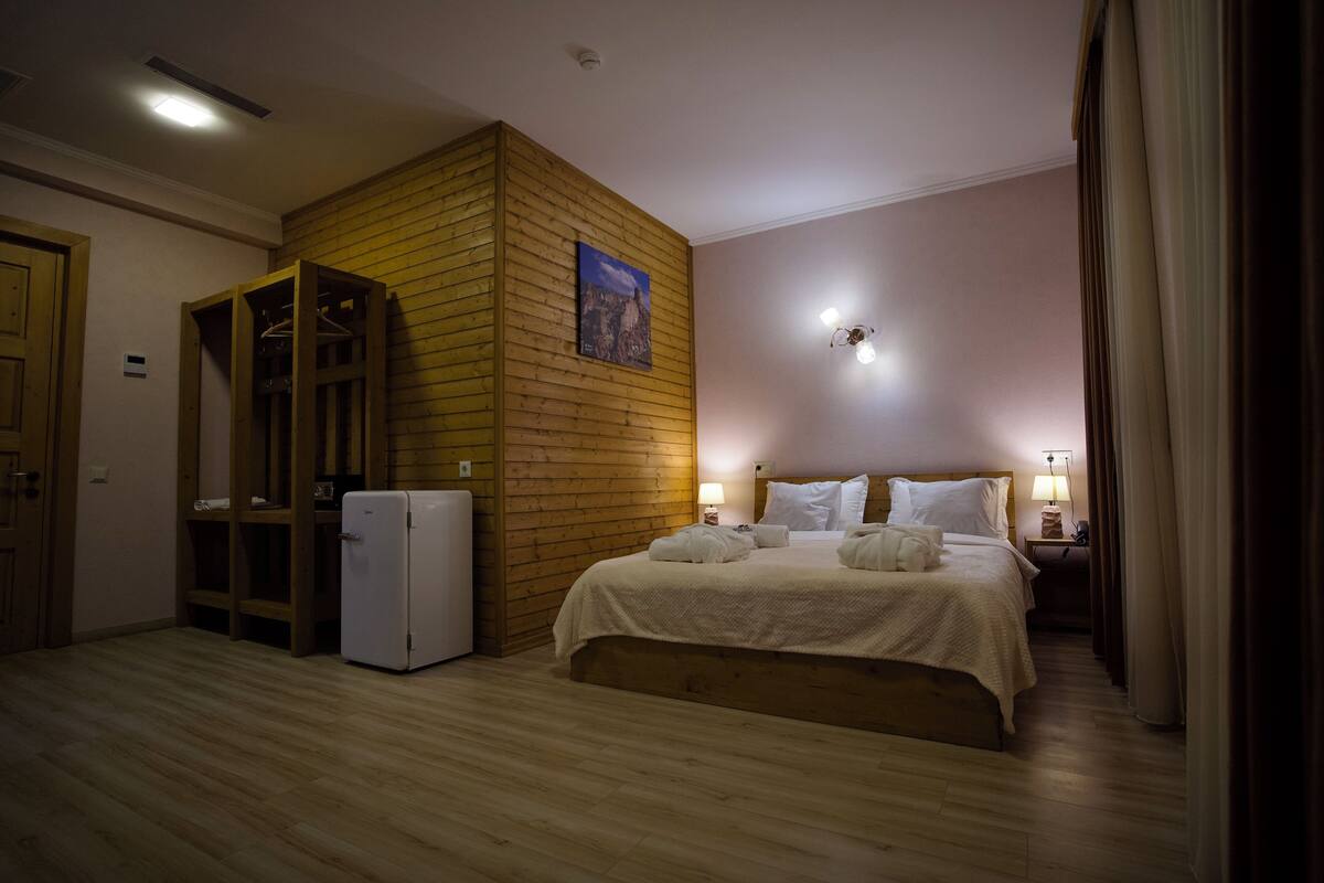 Quarto com guarda-roupa de madeira, cama com roupa de cama branca, frigorífico branco pequeno e luminária fixada na parede.