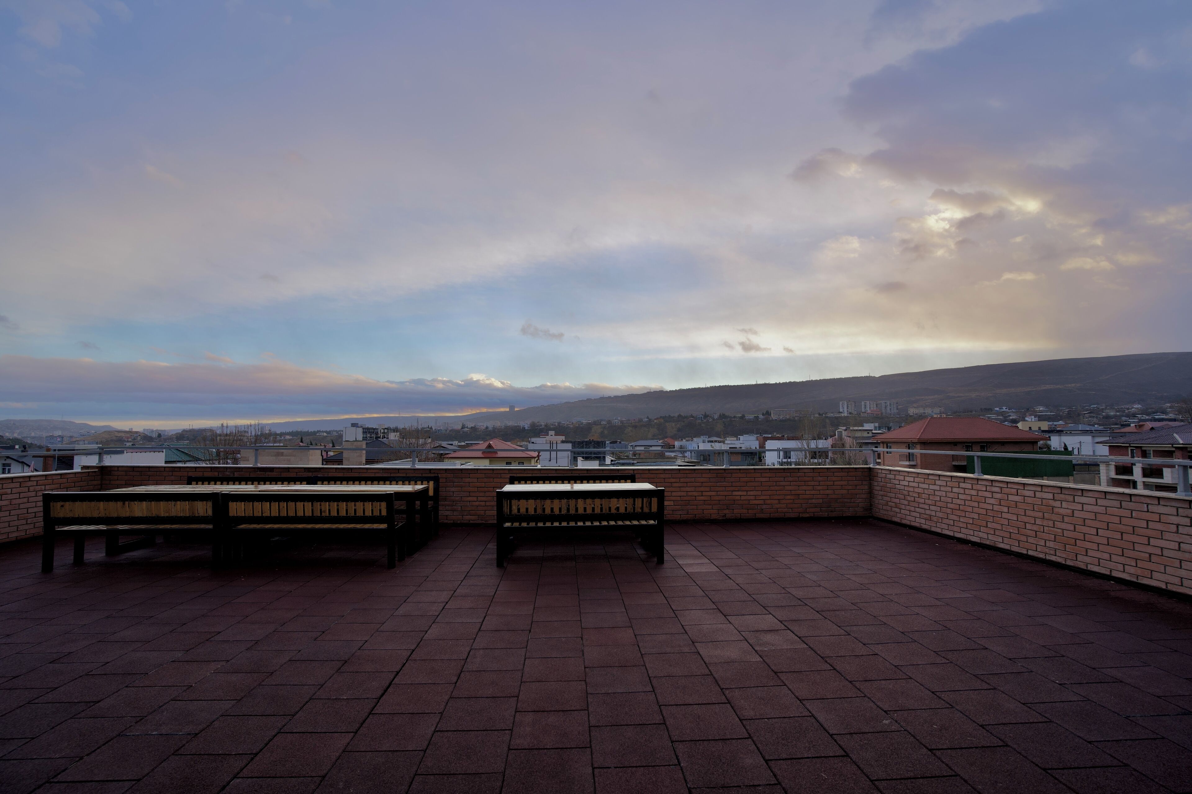A rooftop terrace with two wooden benches and a brick wall, overlooking a cityscape with buildings and hills.