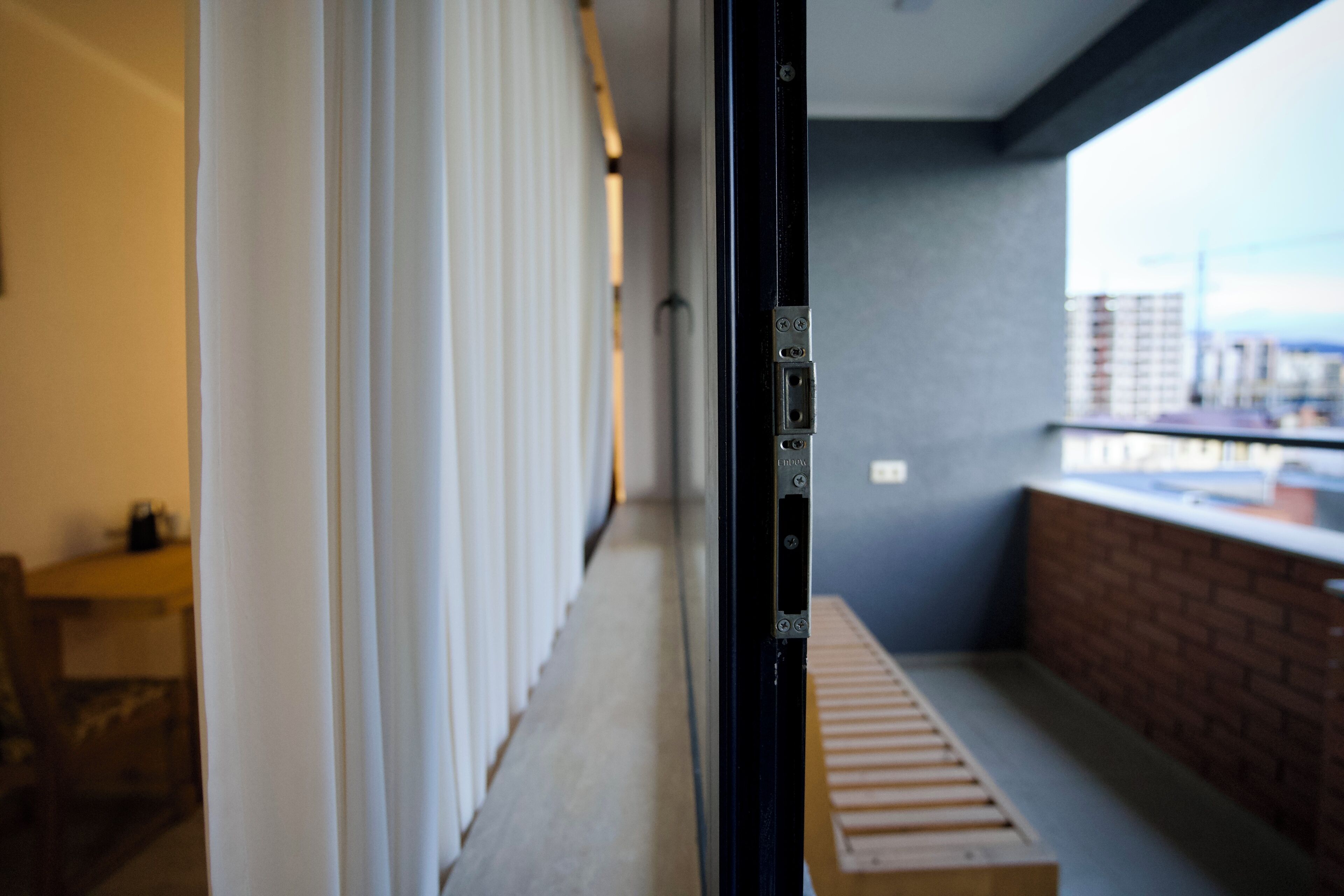 A view from a balcony with a wooden bench, a brick wall, and a window overlooking a cityscape.