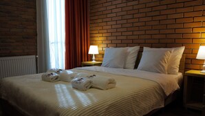 Quarto de hotel com parede de tijolos, cama grande com roupa de cama branca, duas luminárias de cabeceira e uma janela com cortinas.