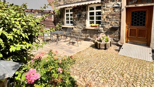 Ein Steinhaus mit Holztür, einer gepflasterten Terrasse und einem Garten mit rosa Blumen.