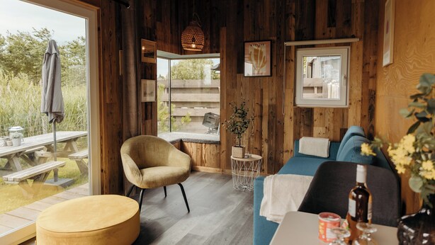 A cozy room with wooden walls, a blue sofa, a round table, and a window overlooking a garden. - Solglimt 2 Pers Holiday Home With Stunning Views (Callantsoog)