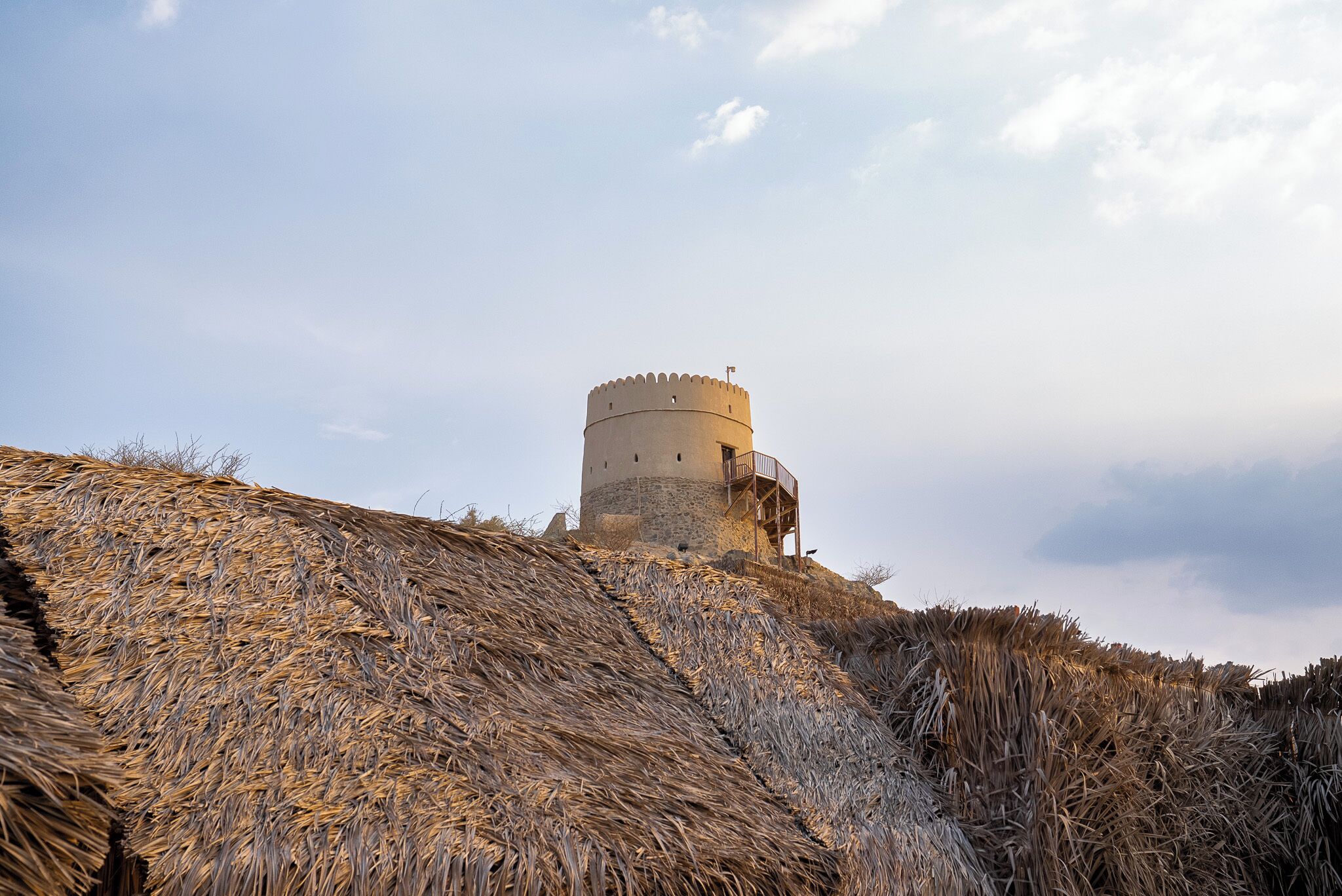 A historic tower with a circular base and a balcony, surrounded by dry grass.