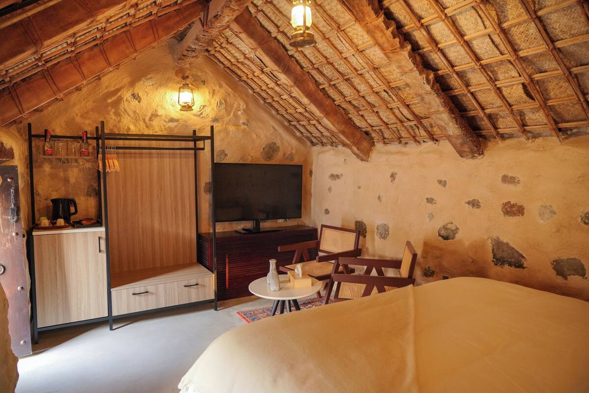 A cozy attic bedroom with a rustic thatched roof, a wooden wardrobe, a television, and a small dining table.
