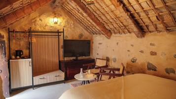A cozy attic bedroom with a rustic thatched roof, a wooden wardrobe, a television, and a small dining table.