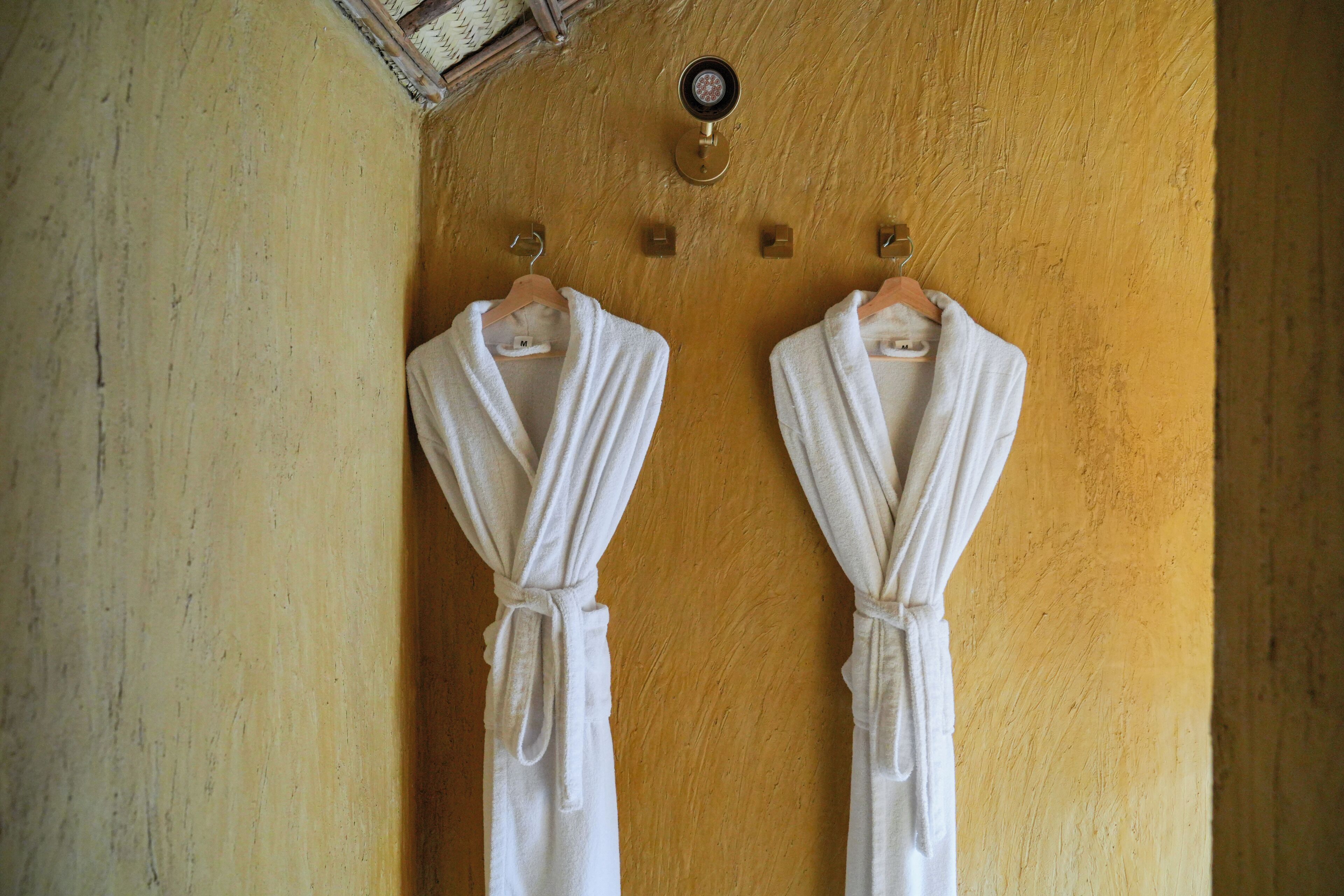 Two white robes hanging on a wooden wall with a small round mirror above them.