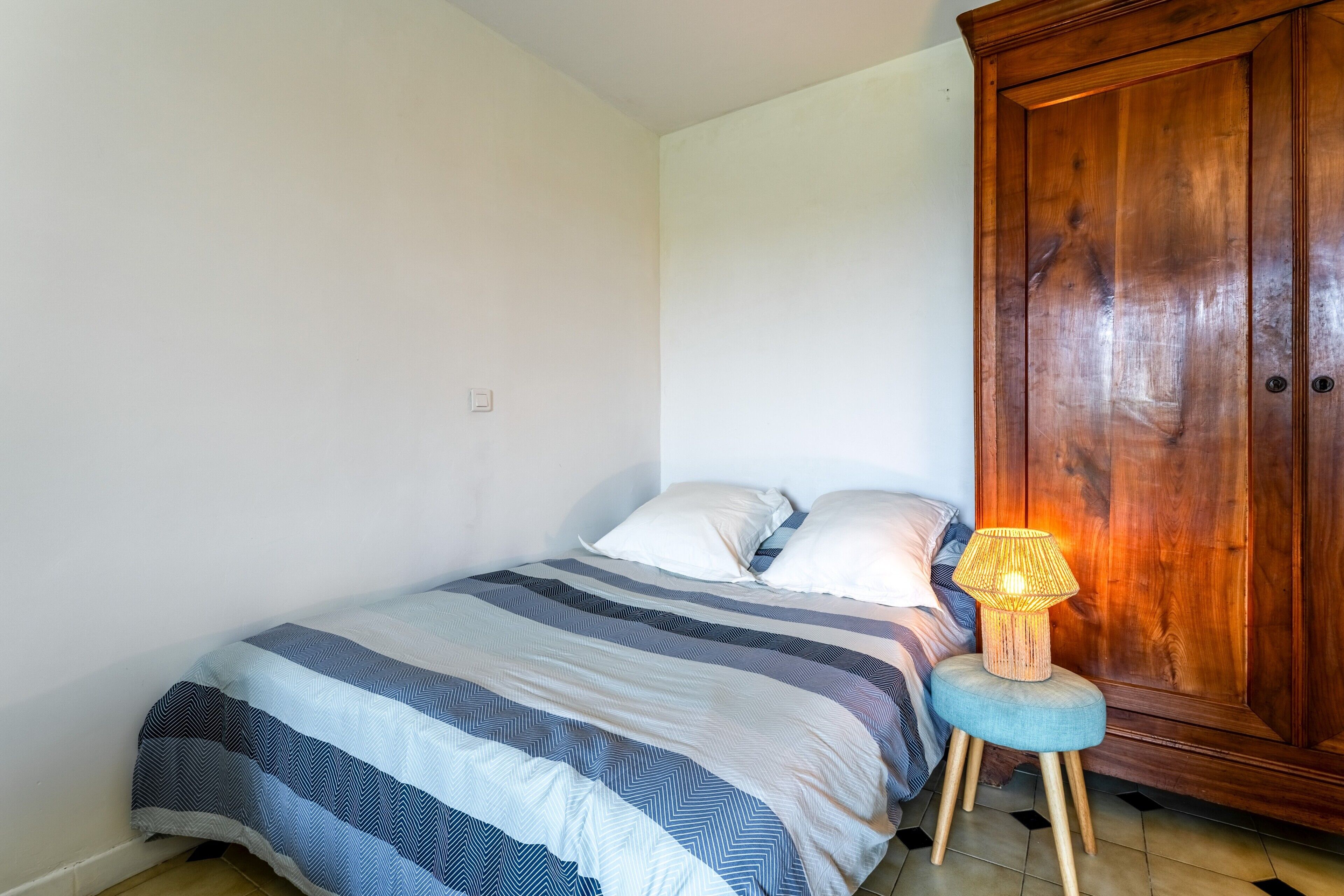Een slaapkamer met een bed, kussens, een bedlampje, een houten garderobe en een klein krukje.