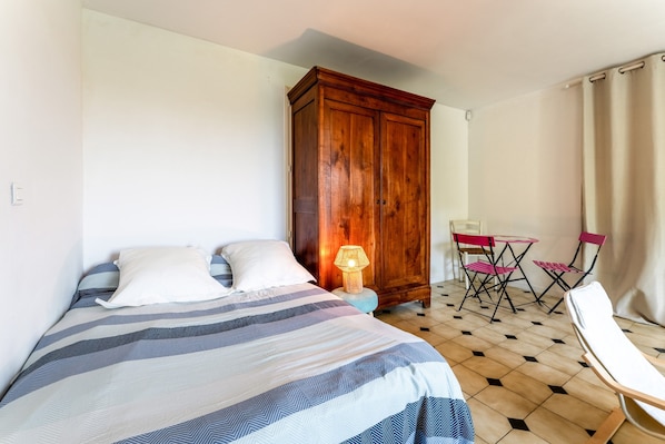 A bedroom with a bed, a wooden wardrobe, a table with chairs, and a lamp.