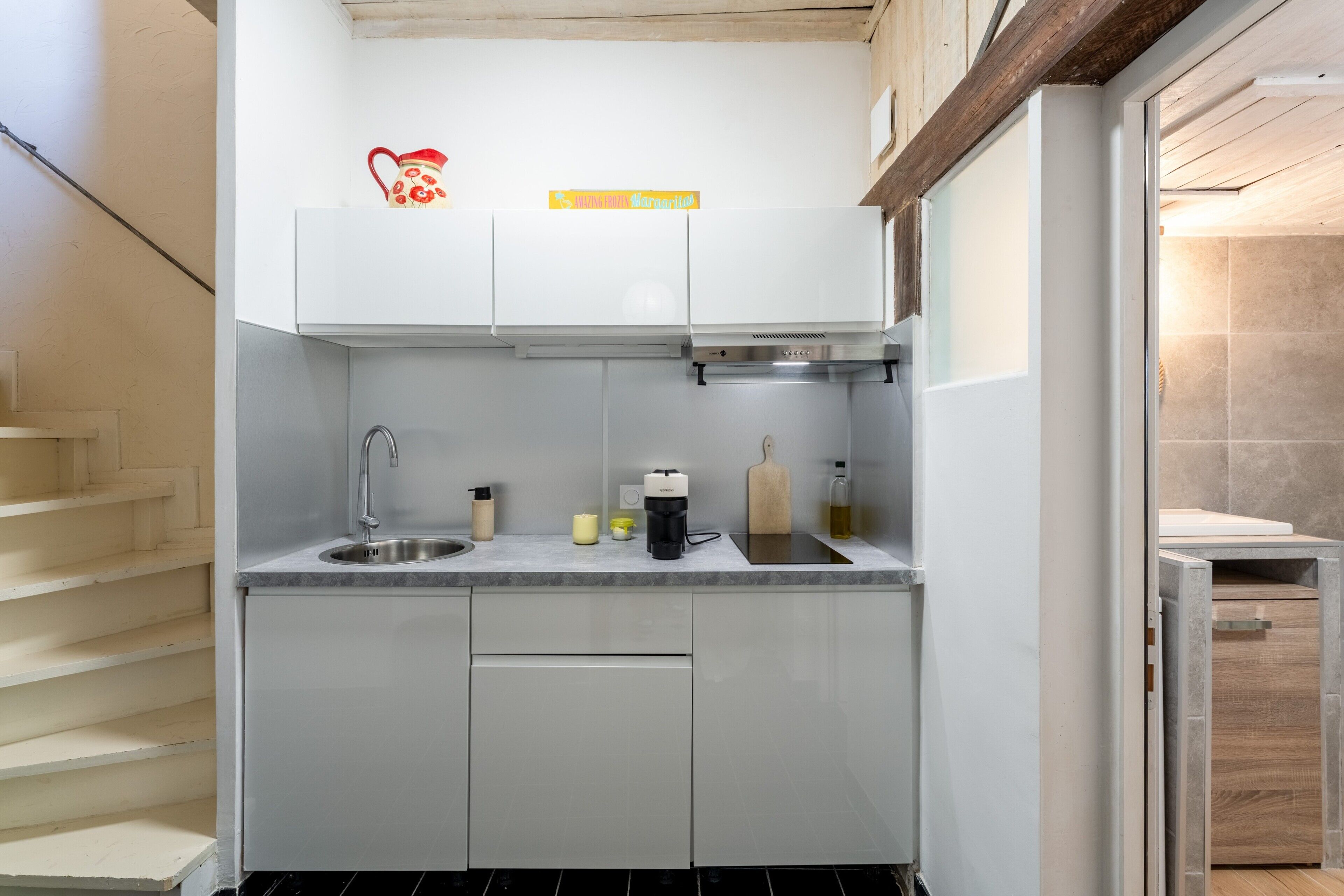 Une cuisine compacte avec des meubles blancs, un évier en acier inoxydable, une machine à café et une planche à découper en bois.