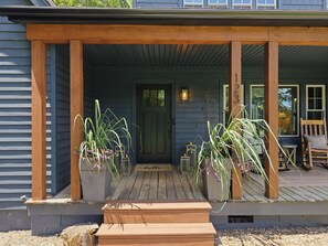 Een houten veranda met een bankje, bloempotten en een deurmat met de tekst 'hello'.