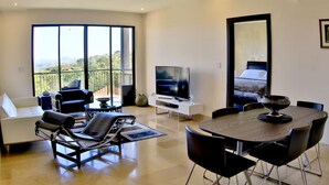 Un salon moderne comprenant un canapé, un fauteuil, une table à manger avec des chaises, un téléviseur et un balcon offrant une vue sur le paysage.