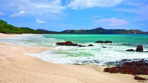 Une plage à l’eau turquoise et limpide, avec un rivage sablonneux et des affleurements rocheux.