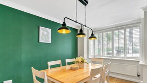 A dining area with a wooden table, white chairs, a green wall, and a window with blinds. - Homely 4 Bedroom House Addlestone (Addlestone)