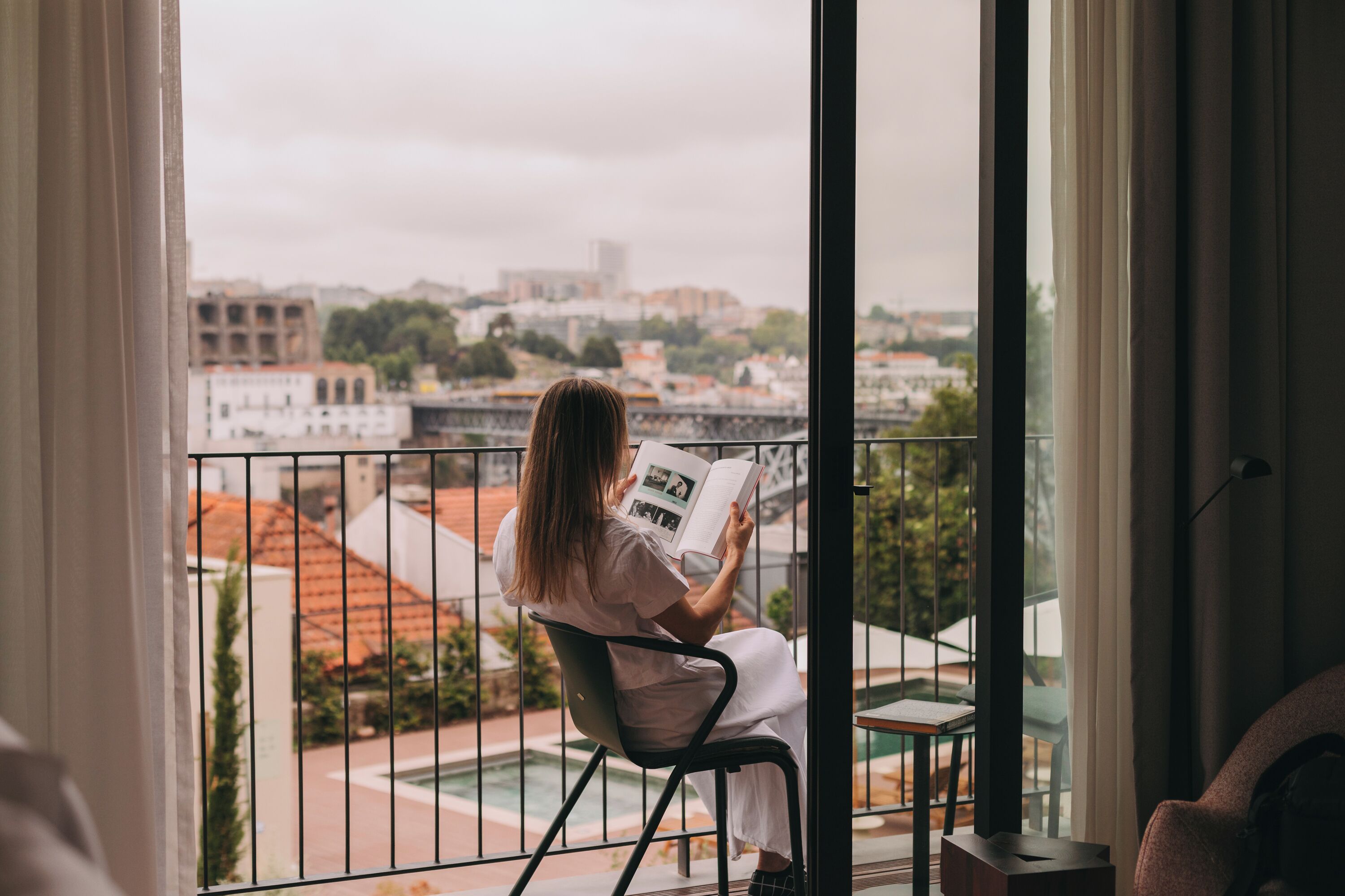 Une personne est assise sur un balcon, un livre à la main, et admire la vue sur la ville.