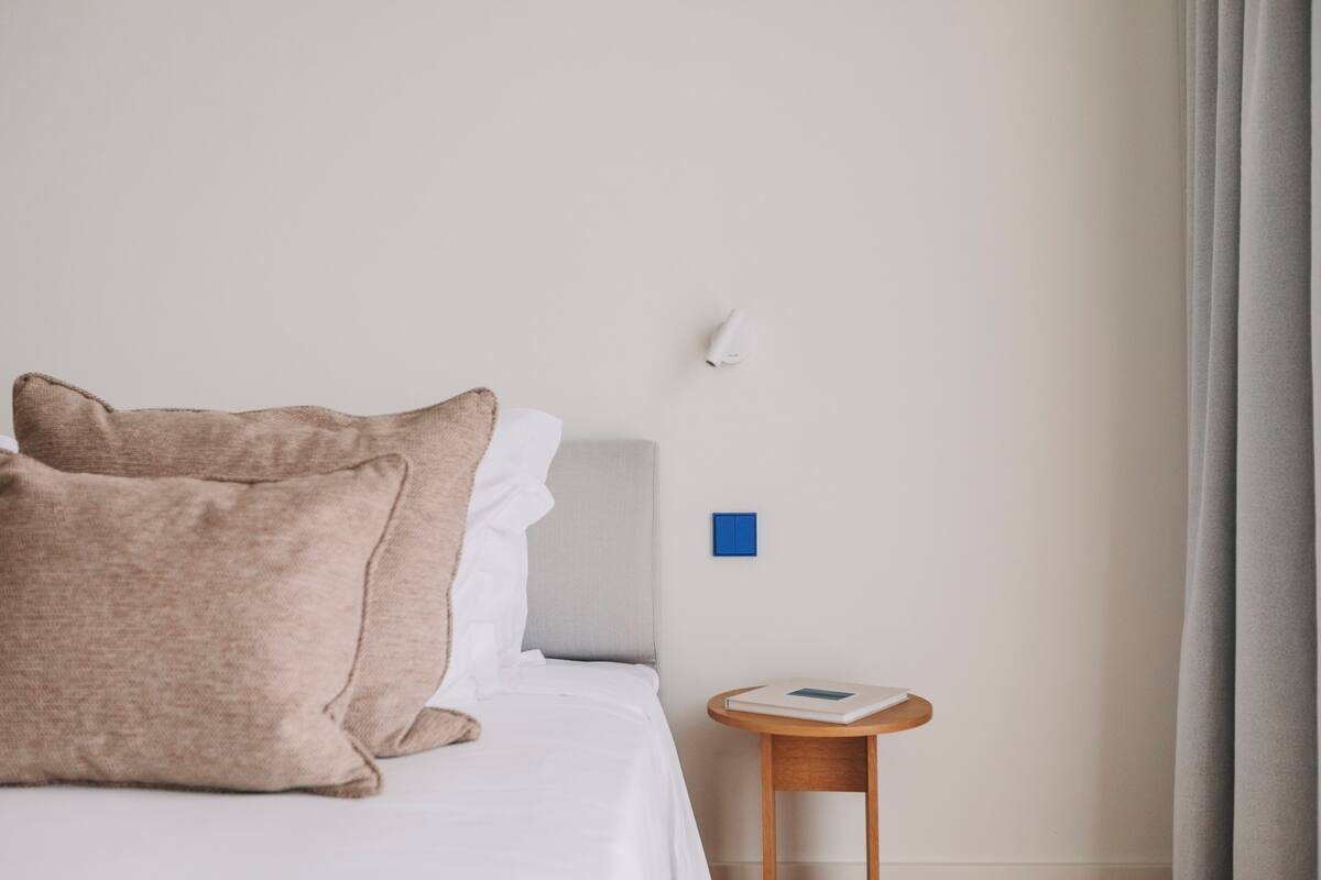 A neatly made bed with two pillows, a small bedside table with a book, a wall-mounted light, and a blue switch.
