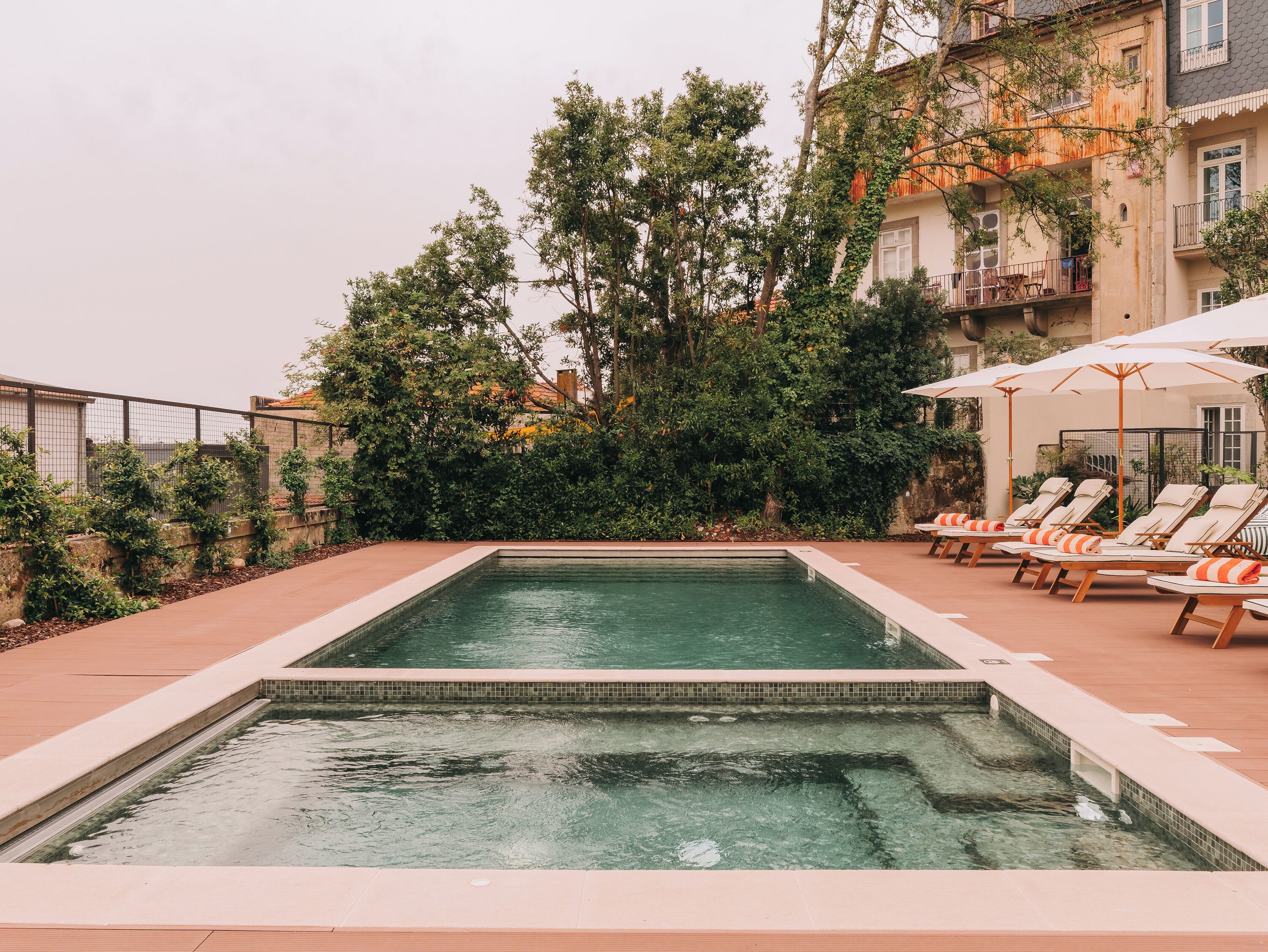 A rectangular pool with a tiled edge, surrounded by a low brick wall and a metal fence. There are lounge chairs with umbrellas and a small table on the right side.