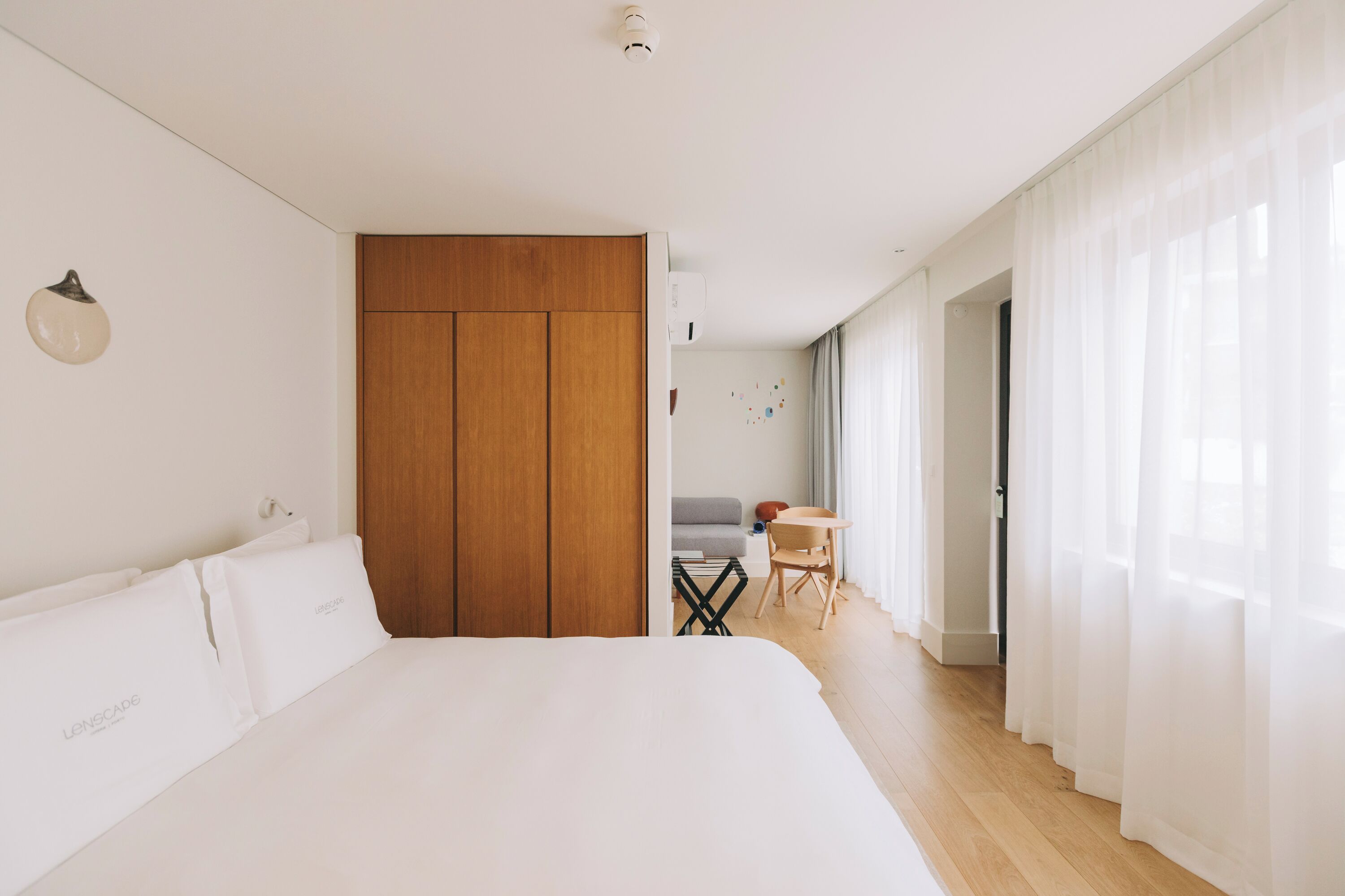 A modern hotel room with a large bed, wooden wardrobe, a small table, and a view of the hallway.