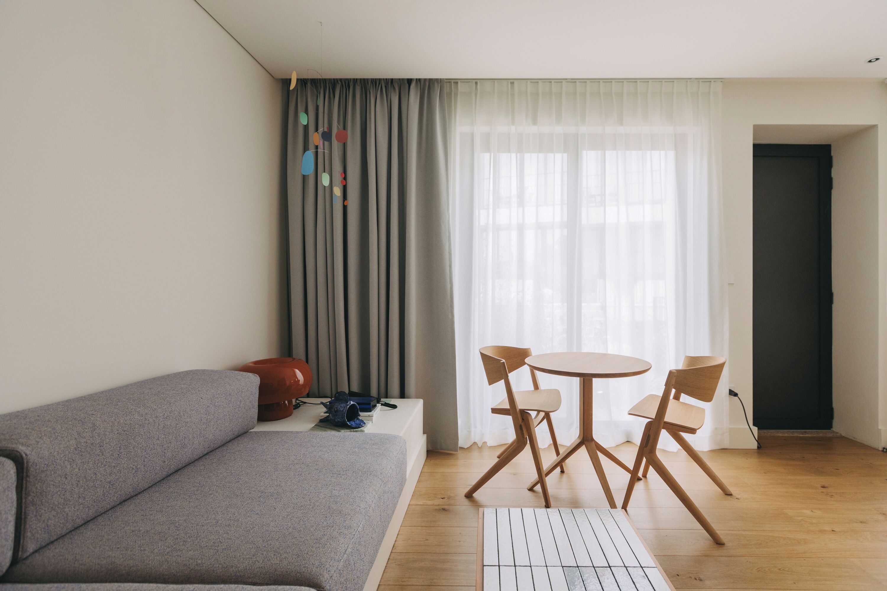 Un salon moderne avec un canapé gris, une table ronde en bois entourée de chaises, un petit tapis et une fenêtre ornée de rideaux légers.