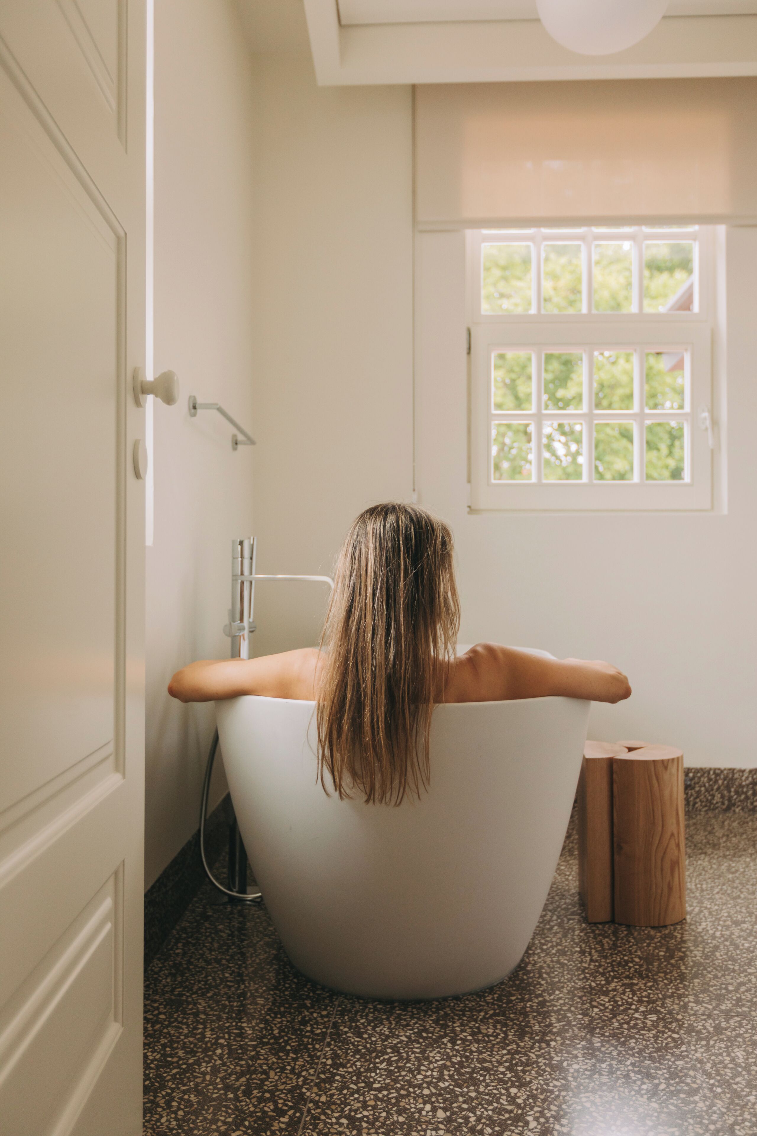 A person in a bathtub with a window above, a wooden stool, and a door.