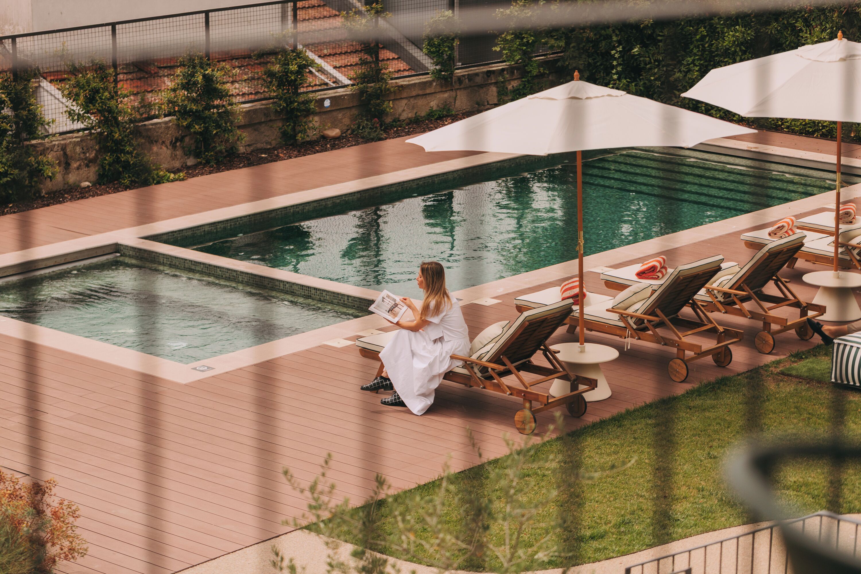 A poolside area with lounge chairs, umbrellas, and a person reading a book.
