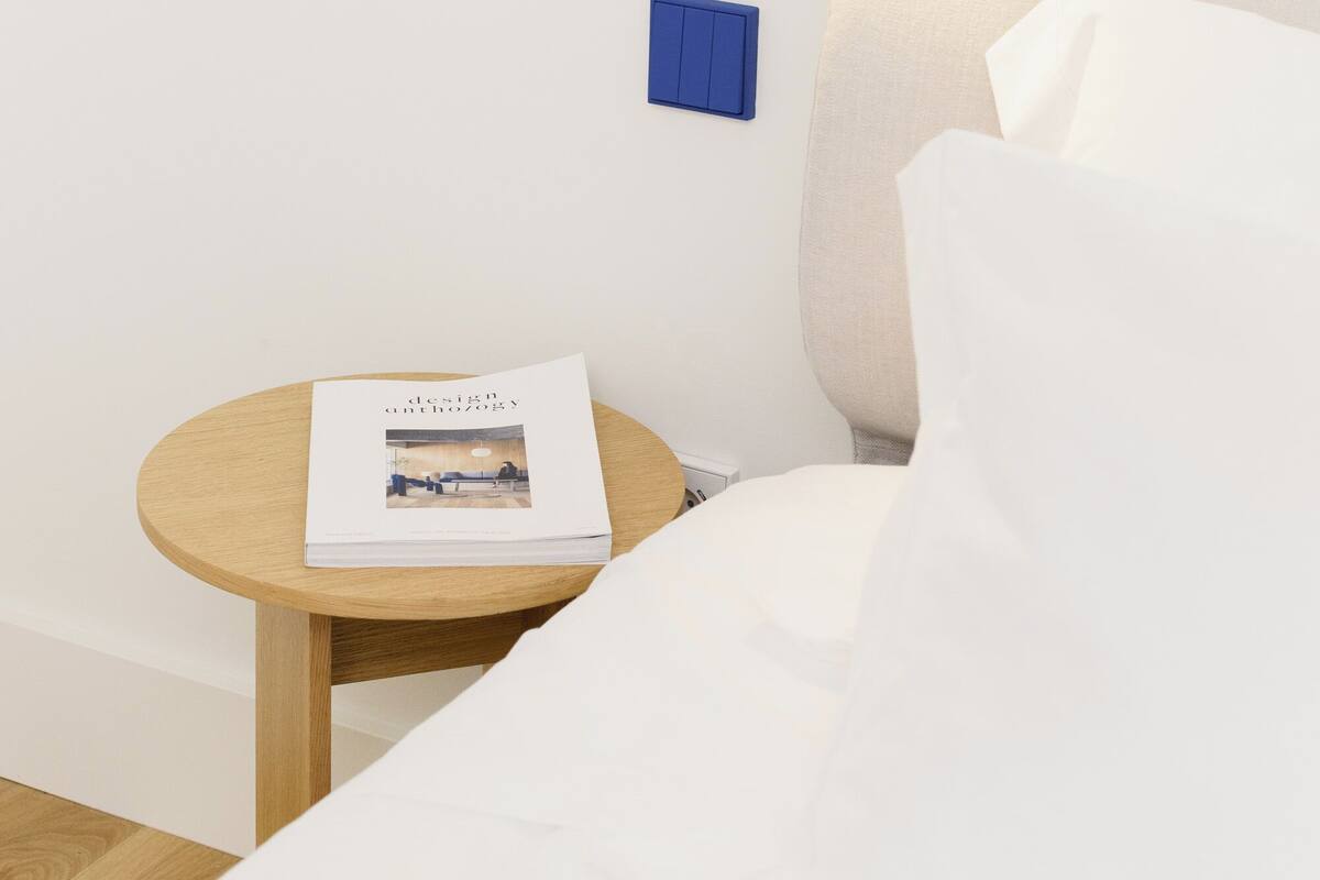 A neatly made bed with white linens, a small round wooden table with a magazine, and a wall-mounted light fixture.