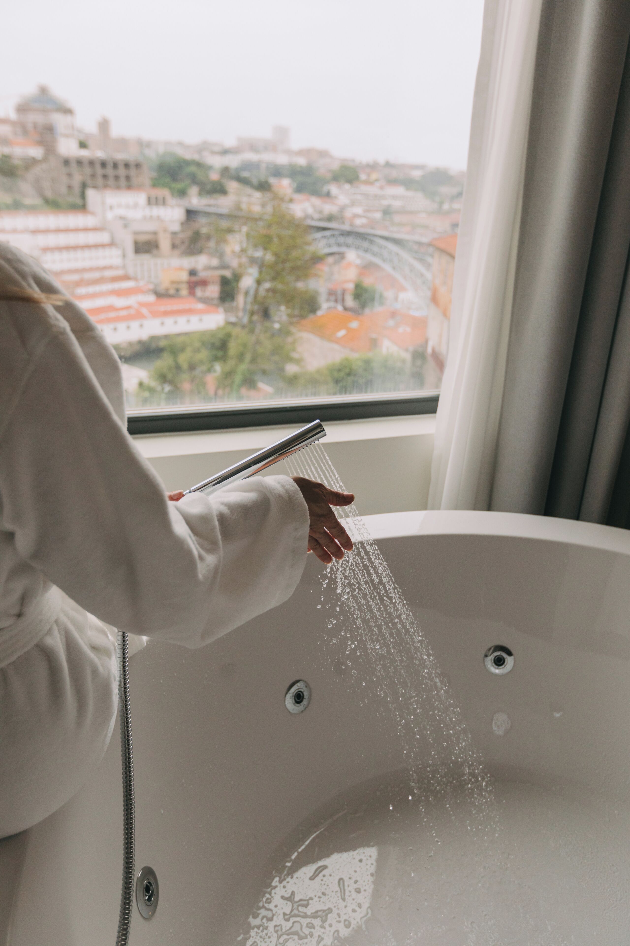 A bathtub with water running, a window with a view of buildings, and a curtain.