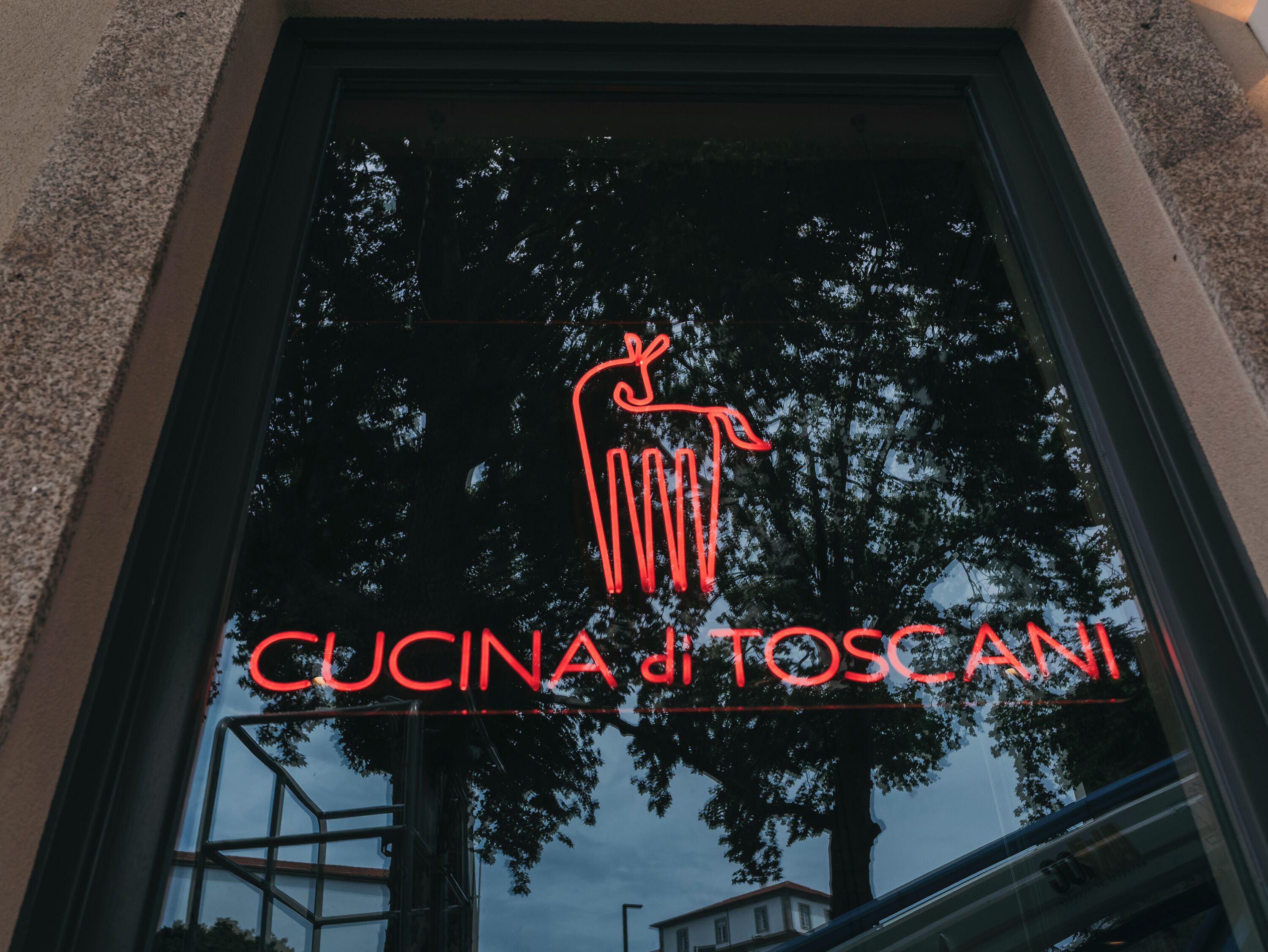 A window with a neon sign reading 'CUCINA di TOSCANI' and a reflection of trees.